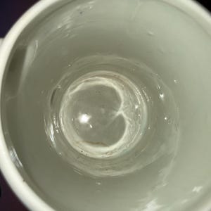 May include: A top-down view of a white ceramic mug with a smooth, glossy interior finish. The bottom of the mug shows a faint, circular brown stain or residue mark, and the cylindrical walls are clean with a reflective, bright white surface.