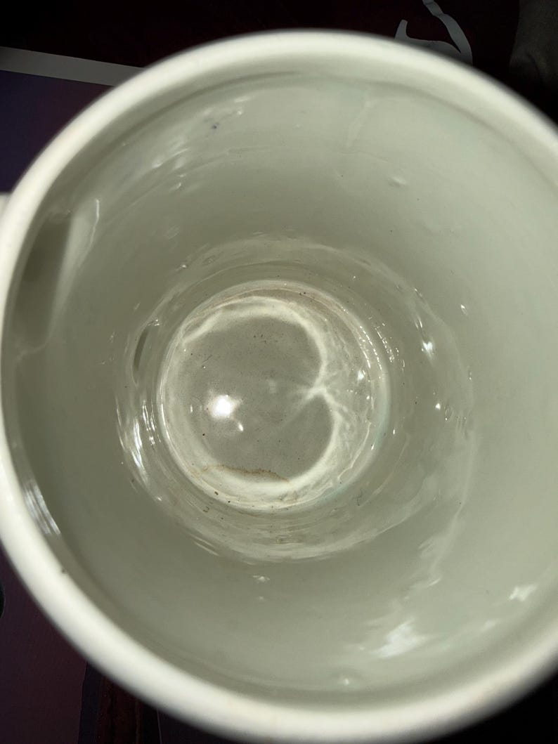 May include: A top-down view of a white ceramic mug with a smooth, glossy interior finish. The bottom of the mug shows a faint, circular brown stain or residue mark, and the cylindrical walls are clean with a reflective, bright white surface.
