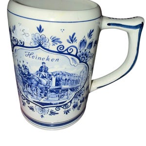 May include: A white ceramic beer mug featuring blue decorative floral patterns and a central illustration of a horse-drawn carriage carrying barrels. The word Heineken is printed in blue script above the carriage scene, and a small price tag is visible.