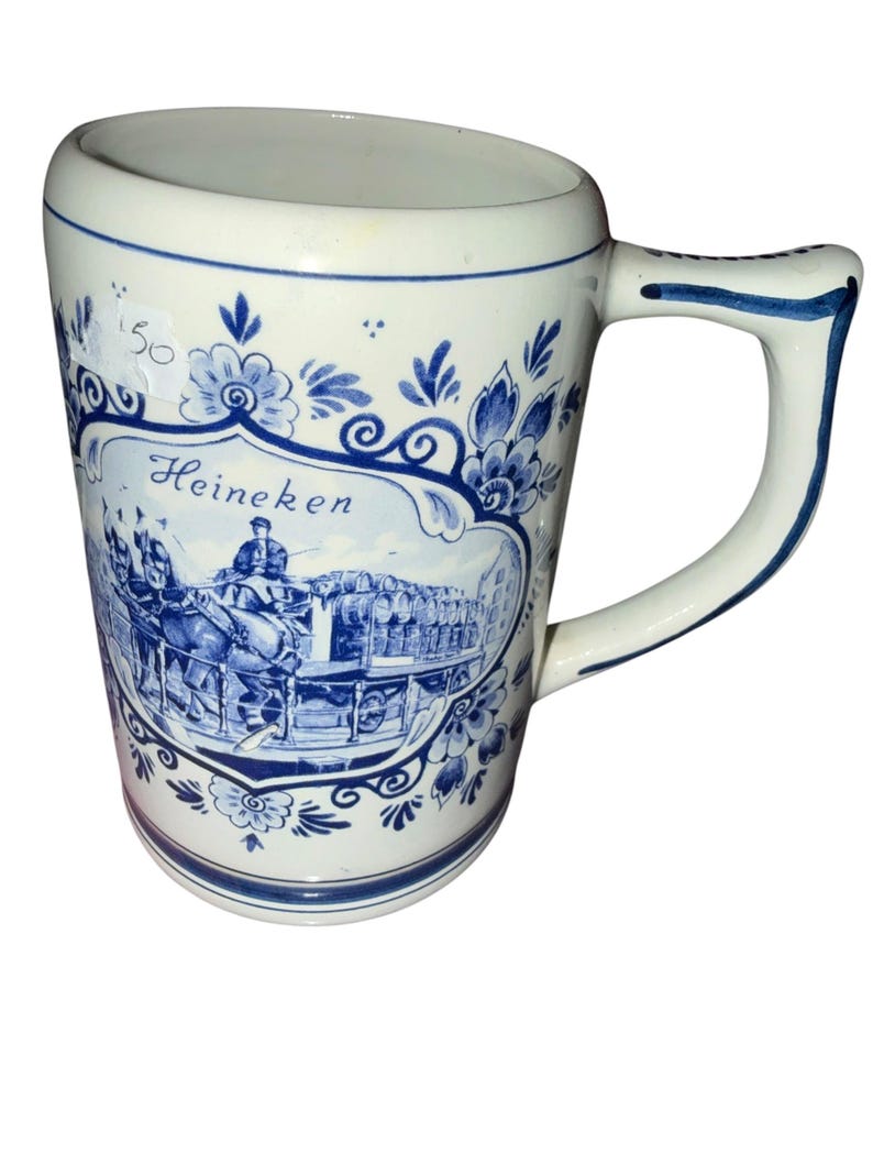 May include: A white ceramic beer mug featuring blue decorative floral patterns and a central illustration of a horse-drawn carriage carrying barrels. The word Heineken is printed in blue script above the carriage scene, and a small price tag is visible.