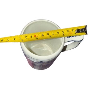 May include: A white ceramic mug with a blue and purple decorative pattern on the exterior. A yellow measuring tape is placed across the top opening, showing a diameter of approximately 3.5 inches. The mug features a handle on the right side.