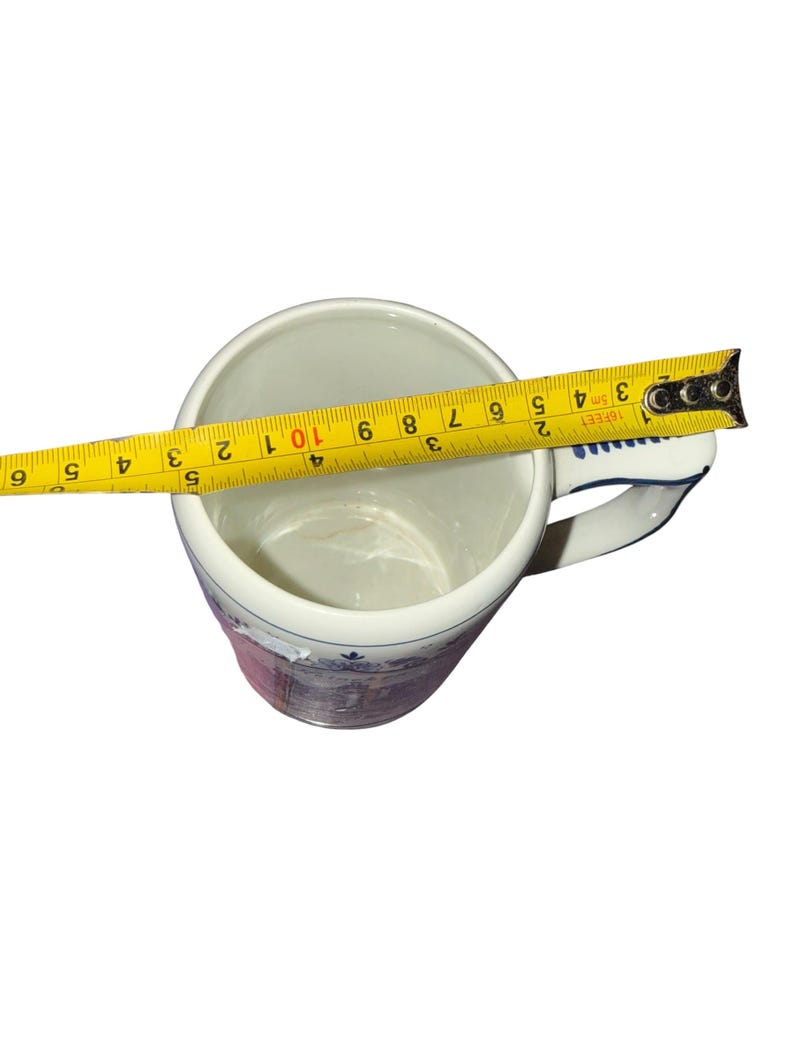 May include: A white ceramic mug with a blue and purple decorative pattern on the exterior. A yellow measuring tape is placed across the top opening, showing a diameter of approximately 3.5 inches. The mug features a handle on the right side.