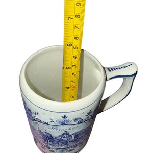 May include: A white ceramic beer mug with blue decorative patterns and the text Heineken. A yellow measuring tape is placed inside the mug, showing a depth of approximately 5.5 inches from the rim to the base of the interior of the vessel.