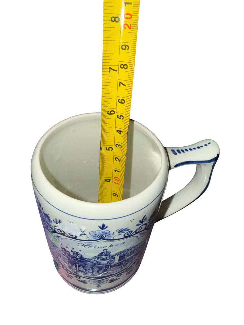 May include: A white ceramic beer mug with blue decorative patterns and the text Heineken. A yellow measuring tape is placed inside the mug, showing a depth of approximately 5.5 inches from the rim to the base of the interior of the vessel.