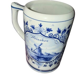 May include: A white ceramic beer mug featuring blue decorative floral patterns and a central illustration of a windmill in a landscape. The word Heineken is printed in blue script above the windmill. The mug has a single handle with blue trim.