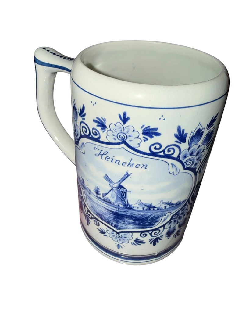 May include: A white ceramic beer mug featuring blue decorative floral patterns and a central illustration of a windmill in a landscape. The word Heineken is printed in blue script above the windmill. The mug has a single handle with blue trim.