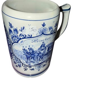 May include: A white ceramic beer mug featuring blue floral patterns and a central illustration of a horse-drawn carriage with the text Heineken. The mug has a sturdy handle and a blue rim accent, with a small white sticker showing the number 50.