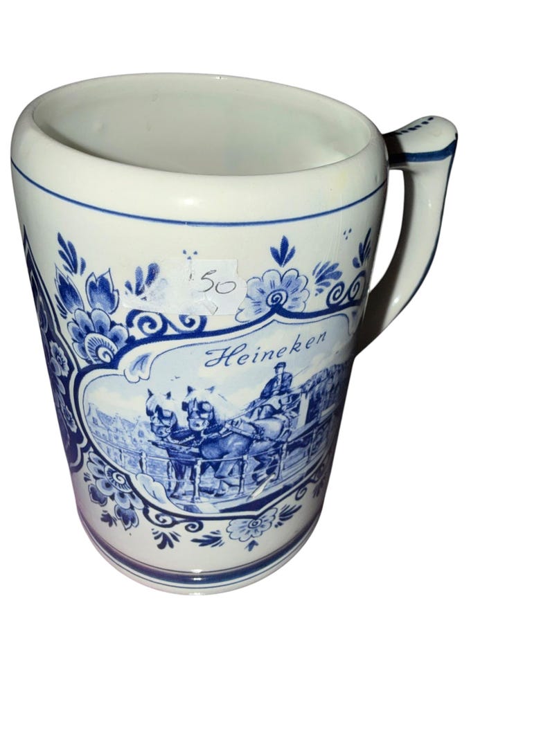 May include: A white ceramic beer mug featuring blue floral patterns and a central illustration of a horse-drawn carriage with the text Heineken. The mug has a sturdy handle and a blue rim accent, with a small white sticker showing the number 50.