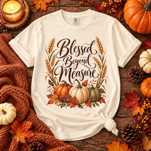 May include: A cream-colored short-sleeve t-shirt featuring a graphic print of three pumpkins, autumn leaves, and wheat stalks. The text "Blessed Beyond Measure" is written in a brown cursive script in the center of the design on the front.