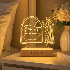 Puede incluir: Una lámpara de acrílico iluminada con un dibujo lineal de una figura y el texto "Fear not FOR I AM WITH YOU ISAIAH 41:10" dentro de un borde arqueado. La lámpara está montada sobre una base de madera rectangular con la marca "Energlyph".