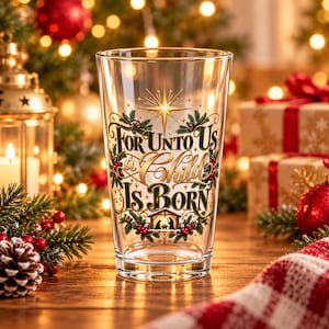 May include: A clear glass tumbler featuring a gold and green printed design with the text For Unto Us a Child Is Born, a star, pine branches, holly berries, and a small nativity scene illustration at the base of the lettering.