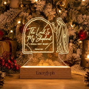 Puede incluir: Un letrero acrílico iluminado con base de madera que presenta el texto The Lord Is My Shepherd I SHALL NOT WANT PSALM 23:1 y una figura de Jesús en arte lineal. La base tiene grabado el nombre Energlyph en fuente oscura sobre la madera clara.