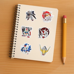 May include: A spiral-bound notebook featuring six distinct Gundam-themed stickers. The designs include various robot figures, a head portrait with Japanese text, and the label RX-78. The stickers are arranged in two columns on the off-white paper.
