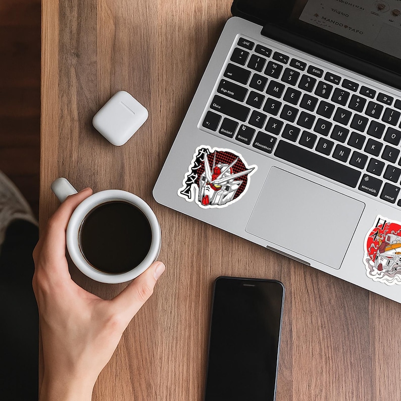 May include: A white vinyl sticker featuring a detailed illustration of a Gundam robot head with red and white accents, set against a black and red dotted circular background, with the Japanese text ガンダム printed vertically in black on the left side.