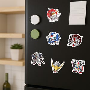 May include: A collection of six vinyl stickers featuring various Gundam robot designs, including full-body mecha figures and head portraits, displayed on a black refrigerator door alongside two circular magnets and a blank white note.