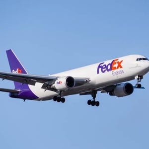 K&ouml;nnte beinhalten: Ein wei&szlig;es FedEx Express-Frachtflugzeug mit lila Akzenten und dem FedEx-Logo in Lila und Rot vor einem klaren blauen Himmel. Das Flugzeug hat zwei Triebwerke und befindet sich im Flug.