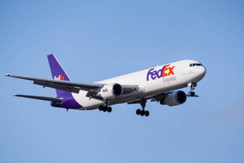 K&ouml;nnte beinhalten: Ein wei&szlig;es FedEx Express-Frachtflugzeug mit lila Akzenten und dem FedEx-Logo in Lila und Rot vor einem klaren blauen Himmel. Das Flugzeug hat zwei Triebwerke und befindet sich im Flug.