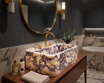 Könnte beinhalten: Ein rechteckiges Aufsatzwaschbecken aus weißem, violett geädertem Marmor mit einer goldenen Armatur mit zwei Griffen. Das Waschbecken steht auf einem dunklen Holzwaschtisch, ergänzt durch ein gefaltetes beiges Waffelpikee-Handtuch und zwei kleine Glasflaschen.