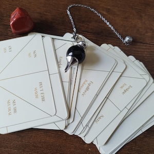 Puede incluir: Un juego de adivinación con un péndulo y una baraja de cartas. El péndulo tiene una piedra negra en forma de lágrima y una cadena plateada. Las cartas son blancas con detalles dorados y diseños geométricos, con frases como "Sí", "No" y "Neutral". Una piedra roja está cerca.