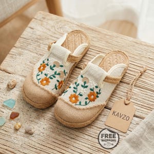 Pode incluir: Um par de sapatos sem atacadores bege com uma textura tecida estilo juta, biqueira arredondada e um padrão floral bordado em laranja, branco e verde. Uma etiqueta de cartão bege com o texto KAVZO está presa na lateral de um sapato.