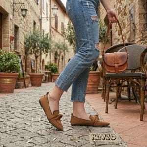 Op de afbeelding: Bruine suède loafers met een strikdetail en een bijpassende bruine leren handtas. De afbeelding toont een persoon die een blauwe jeans draagt en over een geplaveide straat loopt. De merknaam "KAVZO" is onderaan zichtbaar.
