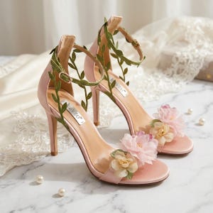 Könnte beinhalten: Ein Paar hellrosa High-Heel-Sandalen aus Satin mit einem Knöchelriemen, der mit grünen Stoffblättern verziert ist. Die Vorderseite jedes Schuhs ist mit geschichteten Stoffblüten in Hellrosa und Cremetönen geschmückt. Der Markenname Kavzo ist sichtbar.