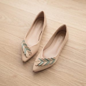 May include: A pair of beige pointed-toe flats featuring a decorative leaf-shaped embellishment on the vamp in shades of sage green and beige. The interior of each shoe is lined in beige and displays the brand name Kavzo printed in black text.