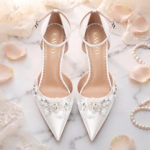 May include: A pair of white pointed-toe high heels with ankle straps, featuring intricate floral beadwork and sequins on the front. The insoles are branded with the text KAVZO in gold lettering, and the shoes have a smooth satin finish.