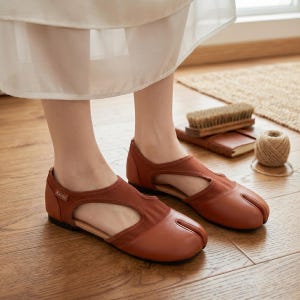 May include: A pair of rust-colored split-toe flats featuring a cutout design on the sides and a small tag labeled Kavzo on the heel. These shoes have a smooth leather-like finish, a flat black sole, and a distinct separation between the big toe and the rest.