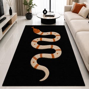 Può includere: Un tappeto rettangolare caratterizzato da uno sfondo nero a tinta unita con un grande motivo a serpente centrato. Il serpente è raffigurato con un motivo sinuoso a forma di S con bande alternate color crema e arancione, inclusa la testa.