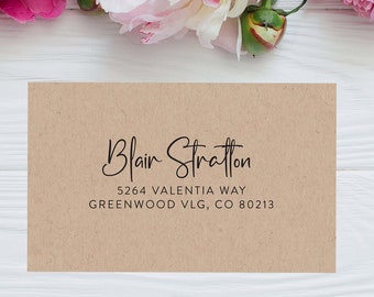 May include: A brown kraft paper envelope with a white background. The envelope has a handwritten address that reads "Blair Stratton, 5264 Valentia Way, Greenwood VLG, CO 80213". The envelope is surrounded by pink and white peonies.