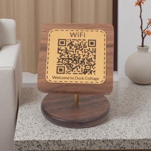 Könnte beinhalten: Ein Ständer aus dunklem Holz mit einem runden Sockel und einer quadratischen oberen Platte, die einen beigefarbenen Einsatz in Lederoptik enthält. Der Einsatz zeigt den Text WiFi, einen QR-Code und den Text Welcome to Duck Cottage.