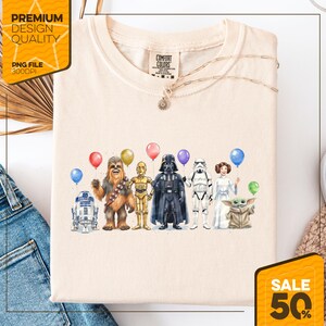 May include: Cream-colored t-shirt featuring a watercolor illustration of Star Wars characters holding balloons. Characters include R2-D2, Chewbacca, C-3PO, Darth Vader, a Stormtrooper, Princess Leia, and Grogu. A necklace is draped across the shirt.