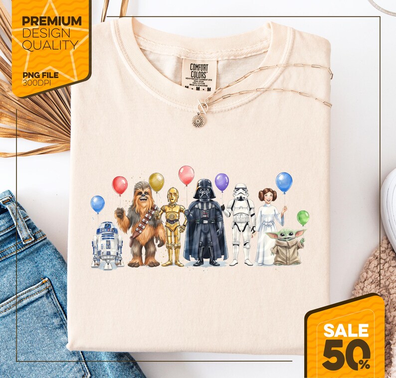 May include: Cream-colored t-shirt featuring a watercolor illustration of Star Wars characters holding balloons. Characters include R2-D2, Chewbacca, C-3PO, Darth Vader, a Stormtrooper, Princess Leia, and Grogu. A necklace is draped across the shirt.
