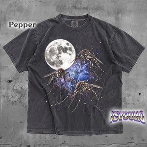 May include: A charcoal grey t-shirt featuring a graphic print of three mosquitoes flying in front of a full moon and a blue nebula. The word Pepper is printed in the upper left corner, and the text VSYOUNG appears in the lower right corner.