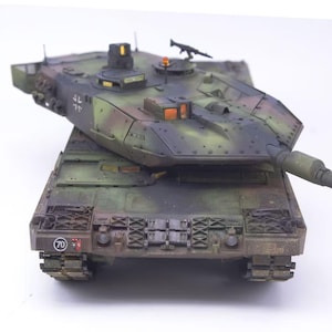 May include: A detailed model tank in a camouflage pattern of green, brown, and black. The tank has a long barrel, a turret with a machine gun, and intricate track details. The number 70 is visible on the front.