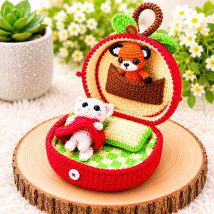 May include: A crocheted red apple-shaped case with a white button closure, containing a white cat wearing a red cape and a green and white checkered pillow. Inside the apple is a crocheted fox in a brown boat.