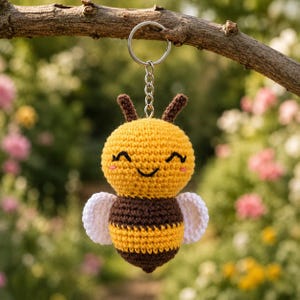 May include: A handmade crochet bee keychain with a smiling face, brown antennae, and white wings. The bee's body is yellow and brown striped. The keychain is hanging from a silver ring and chain, perfect for attaching to keys or a bag.