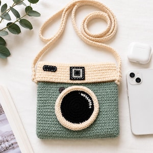 Pode incluir: Uma bolsa tiracolo de crochê em verde sálvia e creme com formato de câmera vintage. A bolsa apresenta um detalhe de lente circular preta, uma seção superior na cor creme com detalhes retangulares pretos e uma alça longa de crochê combinando.