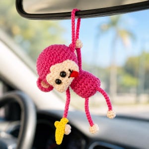 Könnte beinhalten: Ein rosa gehäkelter Affen-Autoanhänger, der an einem Rückspiegel hängt. Der Affe hat ein beiges Gesicht, schwarze Augen und eine rote Zunge. Er hält eine gelbe Banane. Der Anhänger ist handgefertigt.