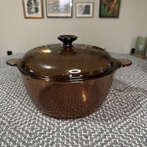May include: A round, translucent amber-colored glass casserole dish with a matching lid. The dish features two integrated side handles and a central knob on the lid. The glass has a smooth, polished finish and a deep brown tint throughout the body.