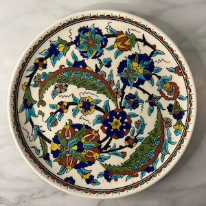 May include: A round, off-white ceramic plate with a detailed floral design. The flowers are primarily blue, yellow, and brown, with green leaves and accents. The rim has a brown border with small, repeating shapes.