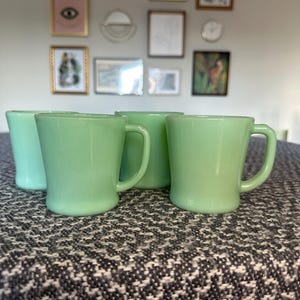 May include: A set of four light green glass mugs with a smooth, semi-translucent finish. Each mug features a rounded body, a wide rim, and a C-shaped handle. The mugs are arranged in a cluster, showcasing their consistent pale jade color and glossy texture.