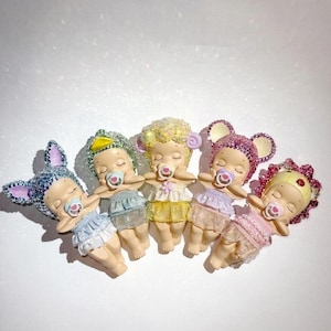 May include: Five small figurines of sleeping babies, each with a pacifier and wearing a different color outfit. The figurines have glittery headwear, including bunny ears, a duck, and mouse ears. The babies are lying down in a row.