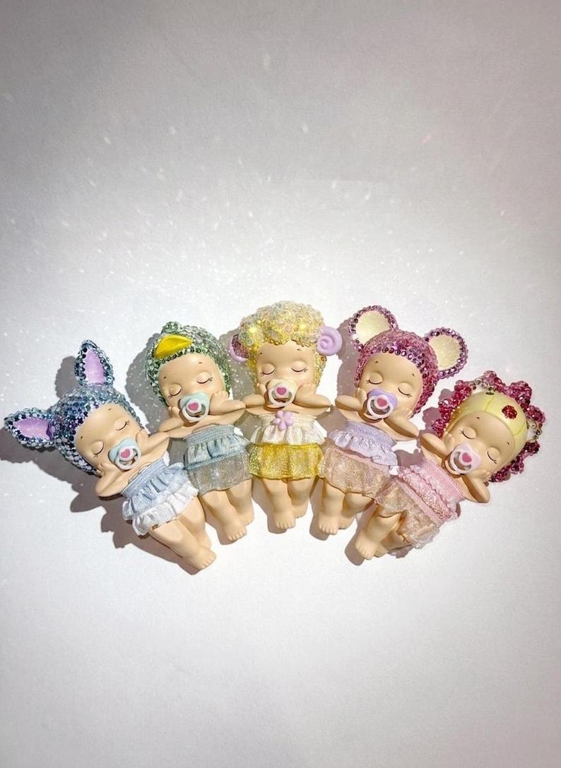 May include: Five small figurines of sleeping babies, each with a pacifier and wearing a different color outfit. The figurines have glittery headwear, including bunny ears, a duck, and mouse ears. The babies are lying down in a row.