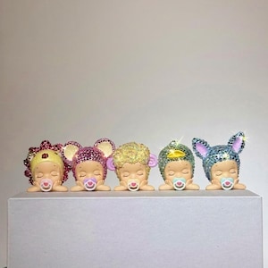 May include: Five figurines of sleeping babies, each with a pacifier and adorned with sparkling headwear. The headwear includes a flower, mouse ears, curly hair, a gem, and bunny ears. The figurines are displayed on a white surface.