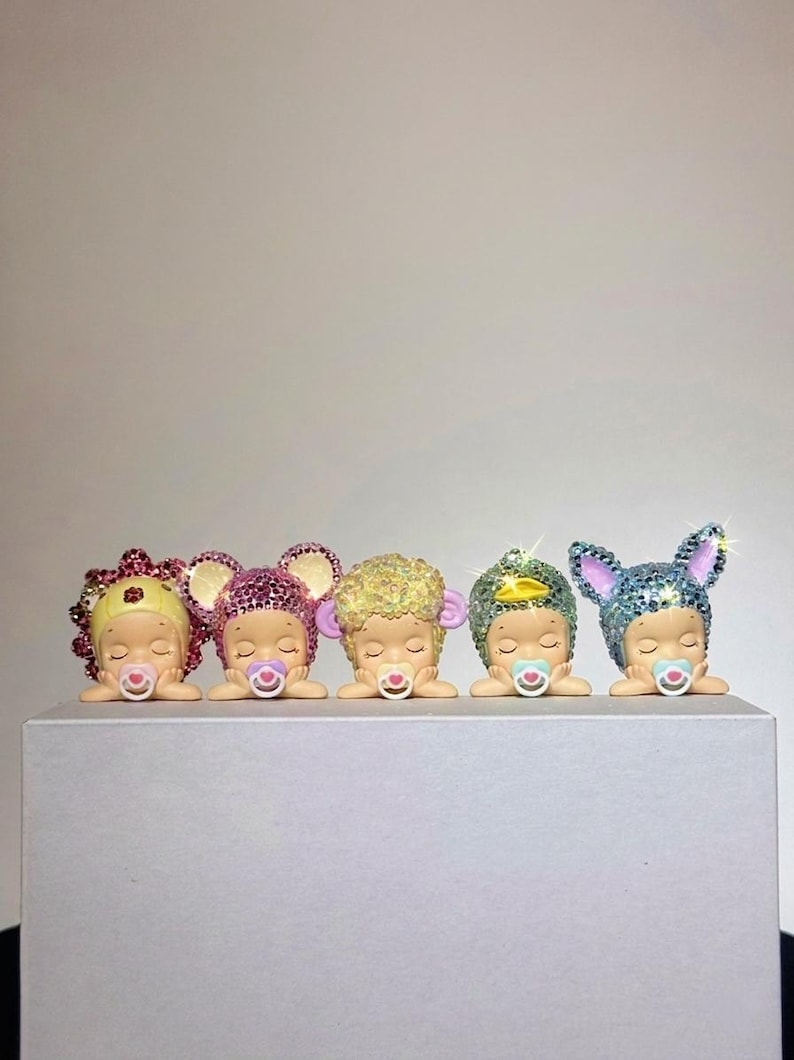 May include: Five figurines of sleeping babies, each with a pacifier and adorned with sparkling headwear. The headwear includes a flower, mouse ears, curly hair, a gem, and bunny ears. The figurines are displayed on a white surface.