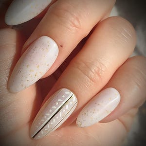 Puede incluir: Primer plano de una mano con uñas artificiales largas en forma de almendra. Las uñas están pintadas de color blanco crema con motas doradas. Una uña presenta un diseño con puntos y líneas blancas y rayas doradas.