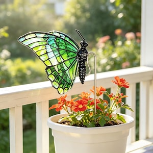 Könnte beinhalten: Ein dekorativer Gartenstecker mit einem Schmetterling aus Buntglas mit leuchtend grünen und gelben Flügeln und einem schwarzen Körper. Der Schmetterling ist auf einem dünnen Metallstab befestigt, der in einem weißen Kunststofftopf mit orangefarbenen Blüten steckt.