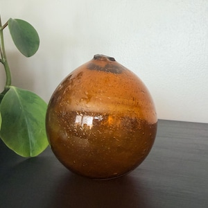 May include: A spherical amber glass vase with a small circular opening at the top. The glass features a translucent, warm brown tone with numerous small air bubbles embedded throughout the material, creating a textured and organic appearance.